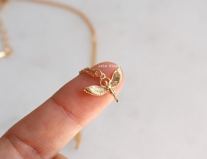 May include: A gold-toned necklace featuring a small pendant shaped like a winged figure with outstretched wings. The pendant is attached to a delicate gold chain. The text "isla flora" is printed in small, light-colored letters above the pendant.