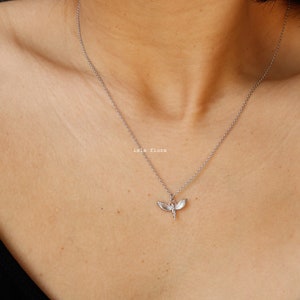 May include: A silver-toned necklace featuring a delicate pendant shaped like a dragonfly with outstretched wings. The pendant hangs from a thin, simple silver-toned chain. The text "isla flora" is printed in small white letters near the pendant.