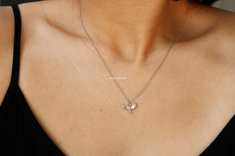 May include: A silver-toned necklace featuring a delicate pendant shaped like a dragonfly with outstretched wings. The pendant hangs from a thin, simple silver-toned chain. The text "isla flora" is printed in small white letters near the pendant.