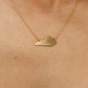May include: A gold necklace with a pendant shaped like the state of Kentucky. The pendant has the words "Isla Flora" engraved on it.