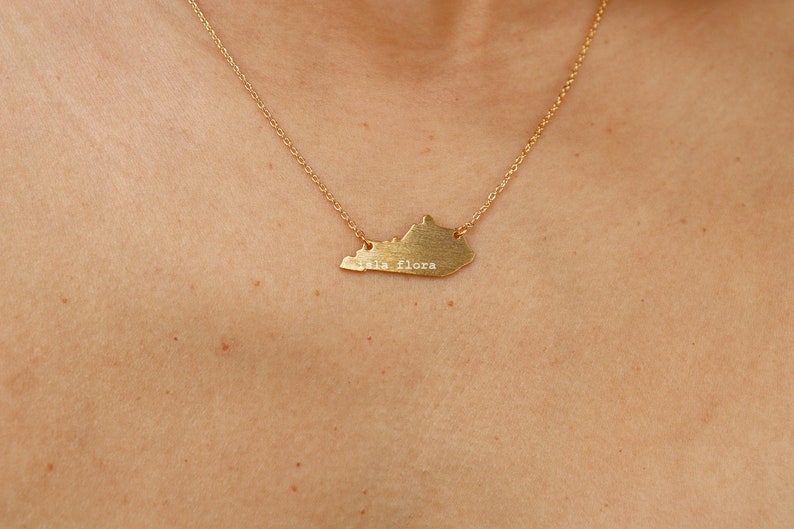 May include: A gold necklace with a pendant shaped like the state of Kentucky. The pendant has the words "Isla Flora" engraved on it.
