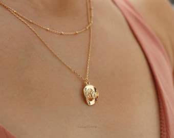 May include: A gold-toned layered necklace featuring two delicate chains. The shorter chain has small bead accents, while the longer chain holds a small, polished gold-toned cowboy hat pendant. The necklace rests against a light pink fabric garment.