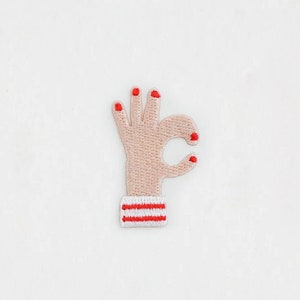 Pode incluir: Patch bordado com um gesto de mão formando o sinal de "OK". A mão é bege claro com unhas vermelhas. O pulso tem uma faixa branca com listras vermelhas. O patch é para roupas ou acessórios.