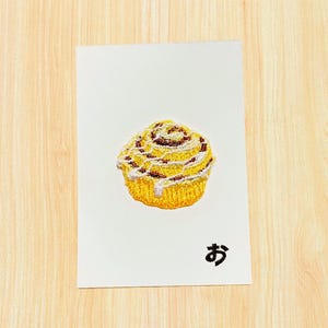 Könnte beinhalten: Eine weiße Karte mit einer detaillierten Illustration einer Zimtschnecke, mit einem goldgelben Boden, braunen Wirbeln und weißem Zuckerguss. Das japanische Zeichen "お" ist unten in Schwarz gedruckt.