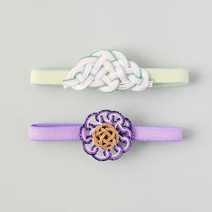 May include: Two hair accessories featuring decorative mizuhiki cord knots. The top accessory has a pale green fabric band with a white and light green woven knot. The bottom accessory has a lavender fabric band with a purple and gold woven knot.