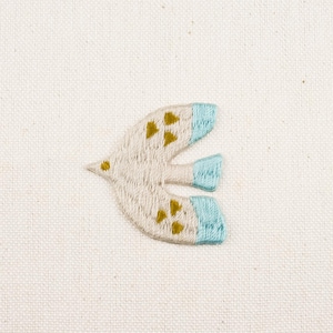 以下が含まれることがあります： 青と金のアクセントが付いた白い刺繍の鳥。鳥は羽を広げて飛んでいる。