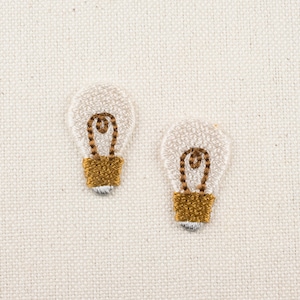 May include: Two white embroidered light bulb shapes with gold accents on a white fabric background.