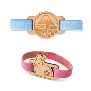 Peut inclure: Deux élastiques à cheveux en bois. L'un présente un motif de montagne et de fleurs avec un élastique bleu clair. L'autre a un motif de lapin et de fleur avec un élastique rose. Les deux sont sur fond blanc.