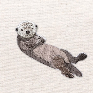 May include: An embroidered patch featuring a sea otter floating on its back. The otter has a light cream-colored head and face with dark brown eyes and nose, transitioning into a textured dark brown and grey body with visible fur stitching.