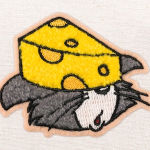 May include: Embroidered patch featuring a cartoon mouse wearing a yellow cheese wedge hat. The mouse has gray fur, white face, and a red open mouth. The patch has a light tan border and is on a white fabric background.