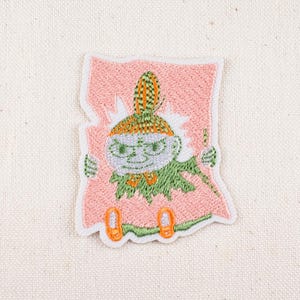 May include: Embroidered patch featuring a whimsical green character with a hat and orange shoes, set against a pink background. The patch has a white border and is likely for clothing or accessories.