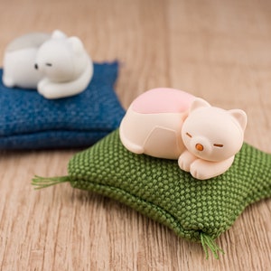 以下が含まれることがあります： 白いセラミック製の小さな猫の置物が2つ、青と緑の布製のクッションの上に寝ています。緑のクッションの上の猫はピンクのお腹をしています。