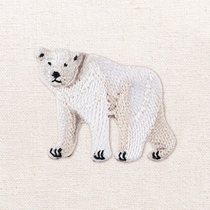 May include: An embroidered patch featuring a white polar bear standing in profile. The bear has detailed white stitching for its fur, with black thread used for its eyes, nose, mouth, and claws. The patch is set against a plain, light-colored fabric.