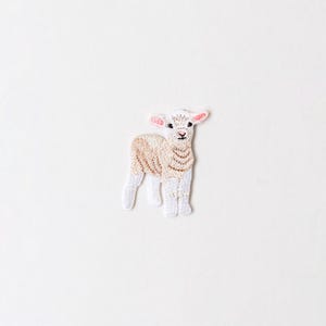 Può includere: Toppa ricamata di un agnello con corpo color crema, zampe bianche e orecchie rosa. L'agnello è posto su uno sfondo bianco. La toppa è probabilmente per abbigliamento o accessori.