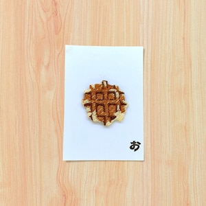 May include: An embroidered waffle patch in brown and tan, on a white card. The card features a Japanese character in black. The background is a light wood grain pattern.