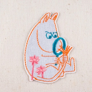May include: Embroidered patch featuring a cartoon character with a magnifying glass. The character is white with orange outlines and details, including the magnifying glass and flowers. The patch is on a light beige fabric.