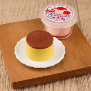Peut inclure: Une gomme en forme de pudding brun et jaune sur une assiette blanche avec un bord festonné. L'assiette est posée sur une surface en bois. On voit une gomme en forme de pudding au lait de fraise rose en arrière-plan.