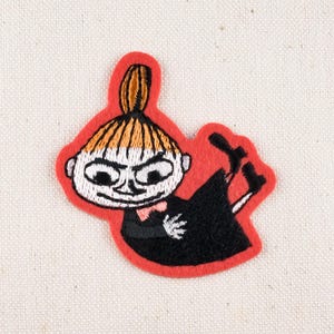 May include: Embroidered patch featuring a cartoon character with a red border. The character has a white face, black eyes, and orange hair. It is wearing a black outfit with a pink bow tie. The patch is approximately 2 inches wide.