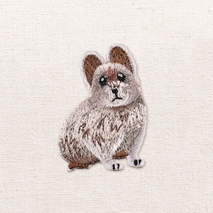 Op de afbeelding: Een geborduurde patch met een kleine, zittende haas met gevlekte bruine, grijze en witte vacht. De haas heeft rechtopstaande oren met een bruine binnenkant, donkere ogen en een kleine neus, weergegeven in gedetailleerd borduurwerk op een lichte stof.