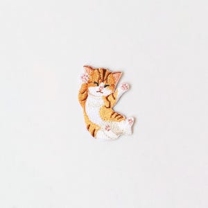 May include: A small embroidered patch featuring a cartoon orange and white cat with closed eyes, lying on its back with its paws in the air.