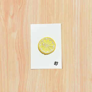 May include: A yellow embroidered lemon slice patch on a white card with Japanese text. The card is centered on a light brown wooden surface. The lemon slice has a detailed design.
