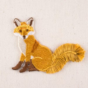 May include: Embroidered patch featuring a sitting fox with orange, brown, and white details. The fox has a fluffy tail and is set against a neutral background. This patch is suitable for clothing or accessories.