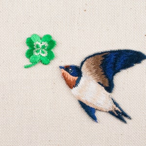 Może przedstawiać: Haftowany znaczek przedstawiający niebiesko-brązową jaskółkę lecącą w kierunku zielonej koniczyny czteroliściennej.