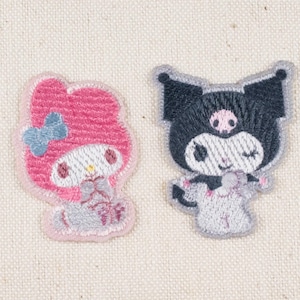 May include: Two embroidered patches featuring cartoon characters. One is pink with a blue bow, the other is black and gray. Both have white faces and are on a beige fabric background. These patches are ideal for decorating clothing or accessories.