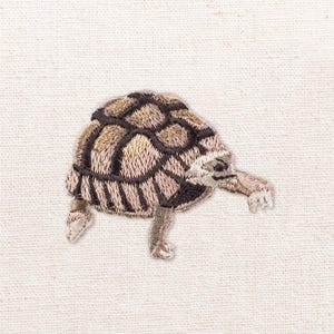 May include: An embroidered patch featuring a tortoise with a textured shell in shades of brown, tan, and black. The tortoise is depicted in a side profile view with its head extended forward and legs visible, showcasing detailed thread work.