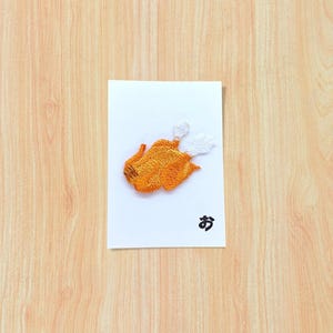 Può includere: Una toppa ricamata raffigurante un pollo arrosto color marrone dorato con punte delle ali bianche, centrata su un cartoncino rettangolare bianco. Il cartoncino mostra il carattere hiragana giapponese nero 'お' nell'angolo in basso a destra.
