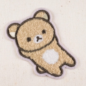 May include: An embroidered patch in the shape of a bear, featuring tan fur, a white belly, and black facial details. It has a light purple border and is set against a cream-coloured fabric. The bear is depicted in a relaxed pose.