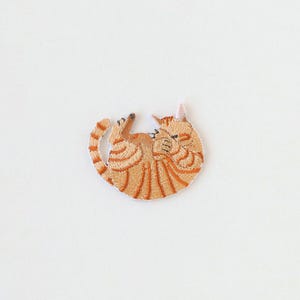 以下が含まれることがあります： 円形に丸まったオレンジ色のトラ猫の刺繍ワッペン。猫にはオレンジ色の縞模様と白い腹部があり、前足を顔の近くに寄せた休息の姿勢で描かれており、細部まで丁寧に刺繍糸で表現されています。