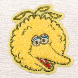 Op de afbeelding: Geborduurde gele patch met een stripfiguur met een grote snavel, grote ogen en een getuft uiterlijk. Het personage heeft een lichtgroene omtrek en drie groene bladeren bovenop. Voor kleding of accessoires.