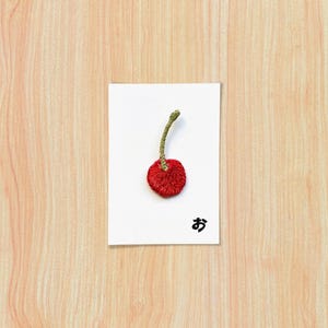 Peut inclure: Une carte blanche avec une broderie de cerise rouge et une tige verte. La cerise est centrée sur la carte, avec un caractère japonais encre noire en dessous. La carte est sur une surface en bois clair.