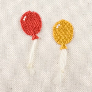 Può includere: Due toppe ricamate a forma di palloncino. Una è rossa, l'altra gialla, ciascuna con un'evidenziazione bianca e un filo bianco. Le toppe sono su uno sfondo di tessuto beige chiaro. Queste toppe sono adatte per decorare abiti o accessori.