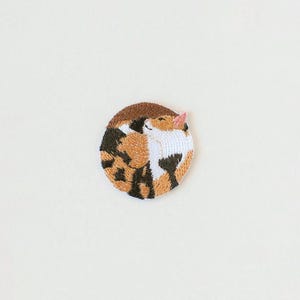 May include: A circular embroidered patch featuring a calico cat curled into a ball. The cat has patches of white, orange, and black fur with a small pink ear visible. The embroidery uses dense thread work to create the feline shape and colour pattern.
