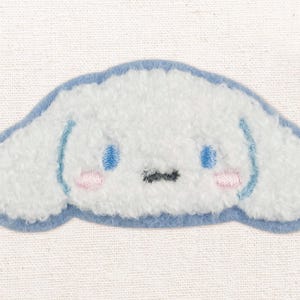 Peut inclure: Écusson brodé blanc et duveteux en forme de tête de personnage de dessin animé. Il présente des contours bleus, des yeux bleus, des joues roses et une petite bouche noire. Le fond est en tissu beige clair.