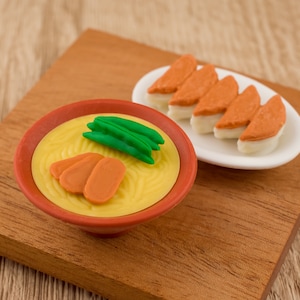 May include: A close-up of two miniature plastic food items on a wooden surface. A bowl of yellow noodles with orange carrot slices and green peas. A white plate with five orange and white fish cakes.