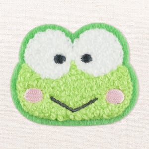 May include: A green frog-shaped embroidered patch featuring large white eyes with black pupils, pink circular cheeks, and a simple black stitched mouth. The texture is raised and fuzzy, with a bright green border outlining the entire shape.