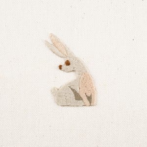 May include: A white embroidered bunny rabbit with brown eyes, sitting on a white fabric background.