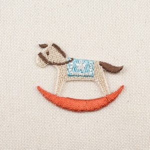 Op de afbeelding: Geborduurd schommelpaard ontwerp in bruin, wit en blauw op een beige stoffen achtergrond.