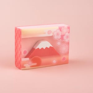 May include: A pink and white soap bar with a design of Mount Fuji and cherry blossoms. The soap is rectangular and has a smooth surface.
