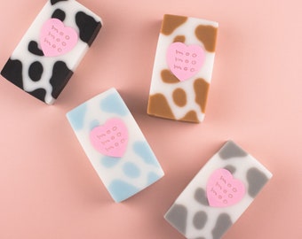 May include: Four bars of soap with a cow print design and a pink heart with the word "moo" printed on it. The soap bars are in black and white, brown and white, blue and white, and gray and white.