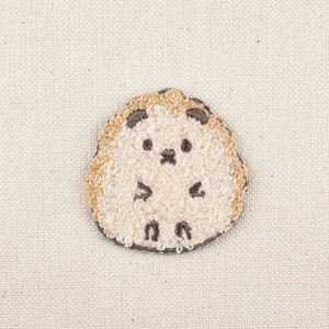 May include: A beige and brown embroidered patch of a cartoon hedgehog with a cute expression.