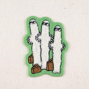 May include: Embroidered patch featuring three white ghost figures with black eyes and small arms, each holding a brown suitcase. The ghosts are set against a green background with a white fabric backdrop.