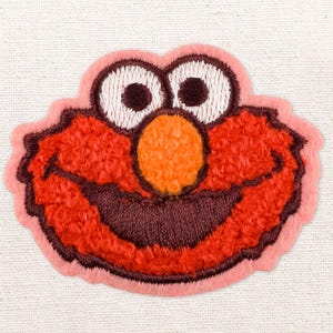 May include: Embroidered patch depicting a cartoon character's face. The character has large white eyes with brown pupils, an orange nose, and a wide, smiling mouth. The face is red with a pink outline, set against a white fabric background.