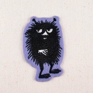 May include: A purple and black embroidered patch featuring a cartoon monster. The monster has a black, spiky body, white eyes, and a white hand. The patch is on a white fabric background.