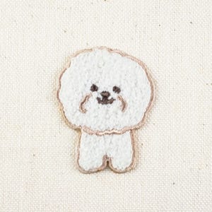 May include: Embroidered patch featuring a cartoon Bichon Frise dog. The dog is white with a fluffy texture, brown eyes, and a brown nose. The patch has a light brown border and is likely for clothing or accessories.