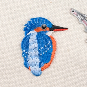 Może przedstawiać: Naszywka haftowana przedstawiająca niebieskiego i pomarańczowego zimorodka z białą rybą obok.