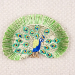May include: A green and blue embroidered peacock with a fringed border. The peacock is depicted with its tail feathers spread out in a fan shape.